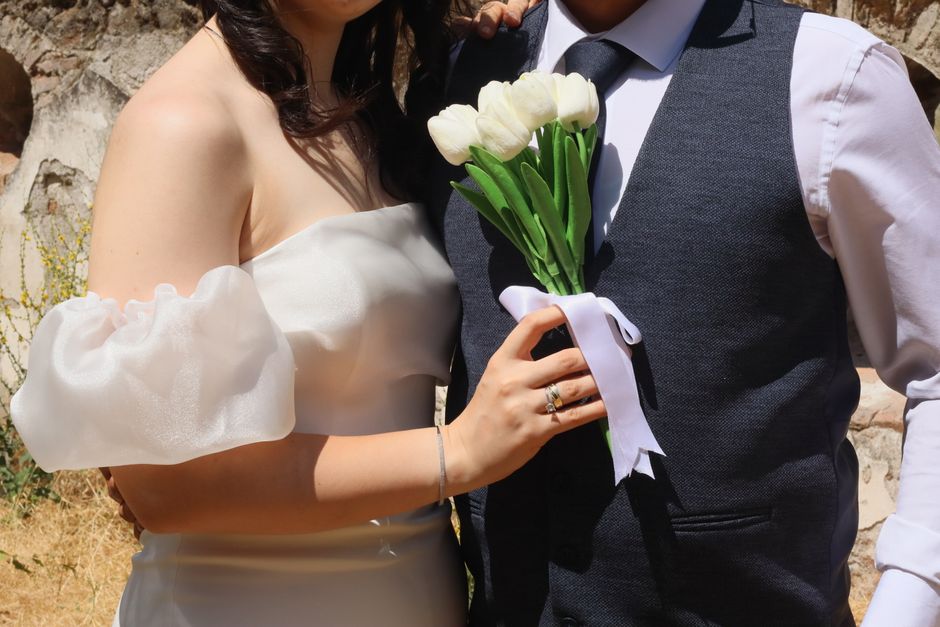 Close-up of a bride and groom at a wedding, with the bride's hand holding a small bouquet of white tulips tied with a white ribbon. The bride wears a strapless white gown with puffed, semi-sheer sleev
