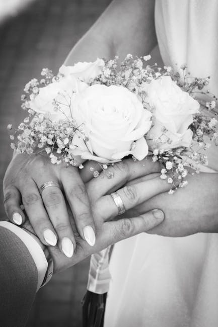 A close-up black-and-white photograph of a bride holding a floral bouquet featuring large, soft white roses surrounded by delicate baby's breath flowers. Her hands, with manicured nails and simple wed