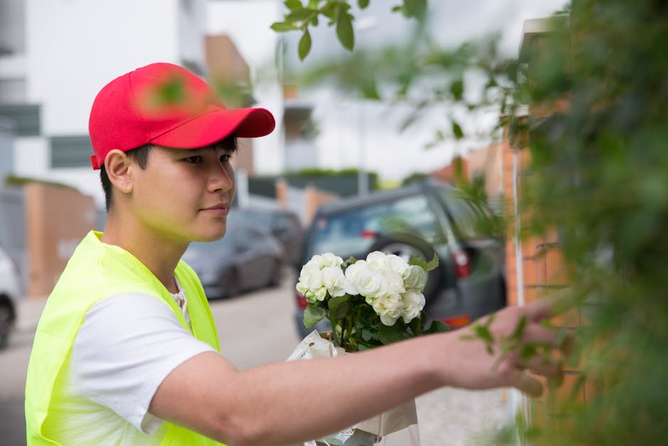 A young male flower delivery courier wearing a red cap and a bright yellow high-visibility vest is seen placing a fresh bouquet of white hydrangeas and roses against a brick wall outdoors. The bouquet