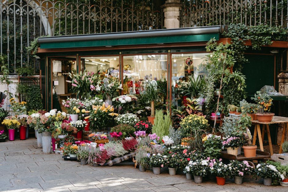 A flower shop storefront with a variety of fresh floral arrangements displayed outside, featuring a mix of colorful blooms including pink, white, yellow, and purple flowers. The arrangement includes r