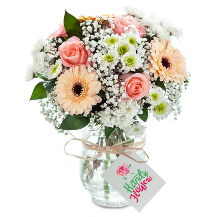 On a gently sunlit surface in a Charlton home, this elegant arrangement of pastel blooms glows with understated sophistication. The bouquet's heart features lush, peachy-pink roses, their petals unfolding in spiral perfection-a striking symbol of care and affection. Tall, peach-coloured gerbera daisies, with vibrant dark centres, rise boldly amid the softer blooms, while the bouquet is made fuller by cushions of dainty white chrysanthemums and clusters of baby's breath, their white florets reflecting the nearby greenery of Maryon Park. Touches of rich, deep green foliage add texture and keep the composition balanced. Each carefully selected stem stands in a clear, subtly patterned glass vase-the water and stems visible, a nod to the freshness and transparency synonymous with local Charlton florists. The vase's neck is circled with rustic twine, from which hangs a crisp white card, the perfect finishing touch for any occasion. This bouquet captures the essence of Charlton's welcoming spirit and is ideal for brightening milestone moments, sending get-well wishes, or creating a warm welcome in any SE7 home.