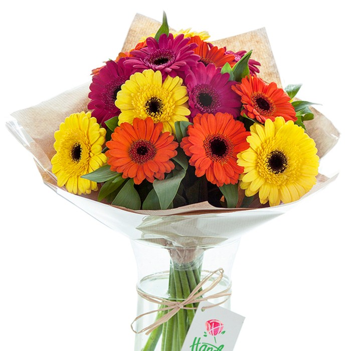 A bouquet bursting with colour and life sits in a clear glass vase, its vivid Gerbera daisies expertly arranged to capture the celebratory energy of Charlton. The arrangement showcases oversized blooms-each with a signature, almost black center ringed by luminous petals in shades of golden yellow, bright orange, and velvety fuchsia-woven effortlessly with sprigs of fresh green foliage to create dynamic depth. The upper part of the bouquet is wrapped in a sheath of rustic light brown kraft paper, lending a homely, artisanal touch reminiscent of mornings at Charlton's farmers' markets, then enveloped by a protective clear cellophane that gently fans out. The flower stems are visible through the vase, partially submerged in water for lasting freshness, neatly bundled and bound with soft raffia. Suspended from the neckline, a small, rectangular white card, adorned with an elegant red rose design and subtle handwritten text, hints at the possibility of a personal message or dedication. The lighting flatters every hue and emphasizes the freshness and care in every detail. This arrangement, evocative of celebratory gatherings in Maryon Park or thoughtful deliveries to local homes, speaks to the artistry and dedication of local Charlton florists, offering a timeless, joyful gesture for birthdays, anniversaries, or moments when only vibrant, hand-tied beauty will do.